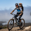 Man riding a mountain bike with a dog on his shoulders against a scenic background
