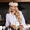 Woman in a kitchen wearing a robe and sleep mask, holding a plate of food and a box labeled 'nodpod'.