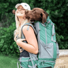 Woman with a green backpack and a dog on her back, surrounded by greenery.