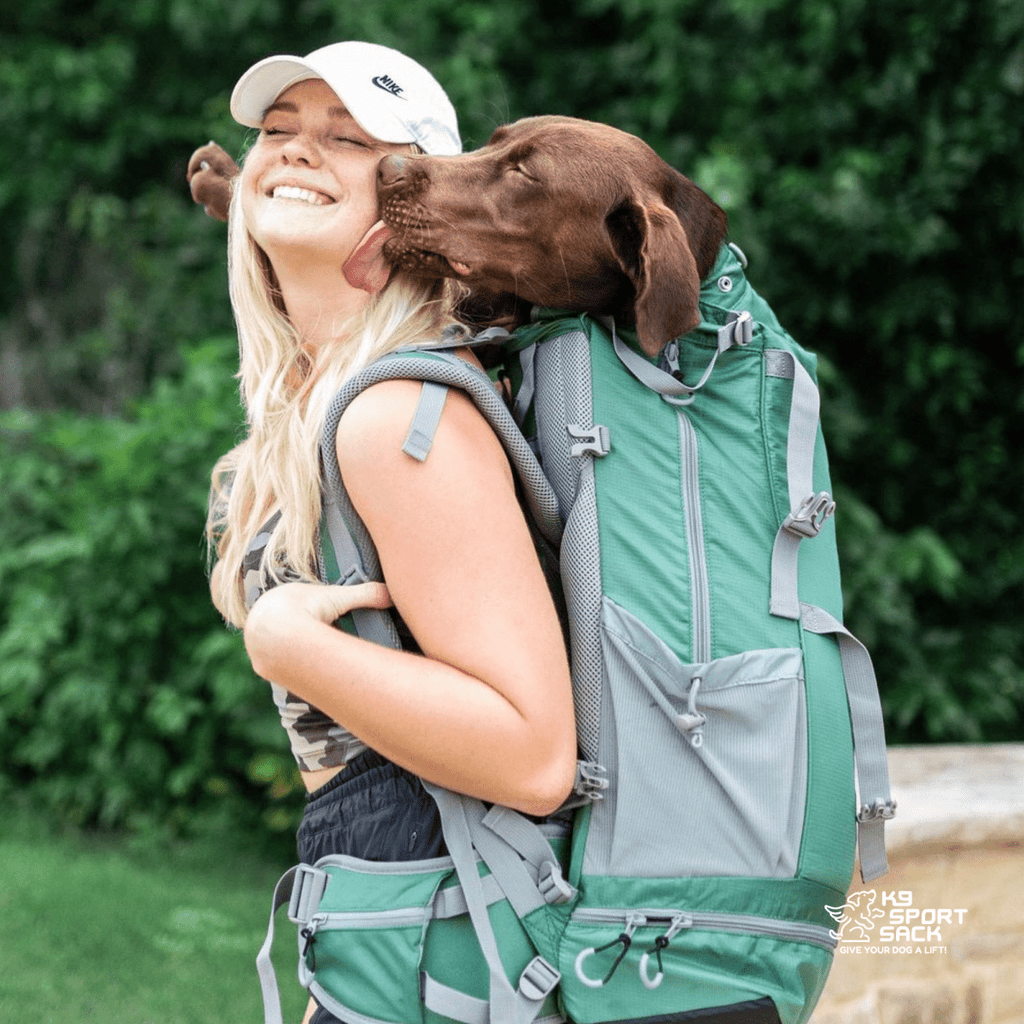 Woman with a green backpack and a dog on her back, surrounded by greenery.