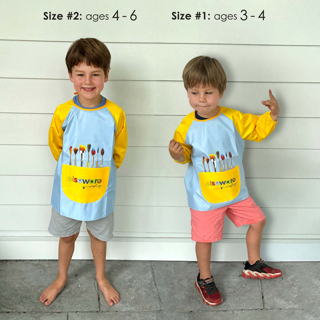 Two children wearing matching aprons with paintbrush pockets against a wooden background.