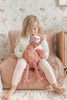 Child sitting on a pink chair holding a plush flamingo toy in a room with floral wallpaper.