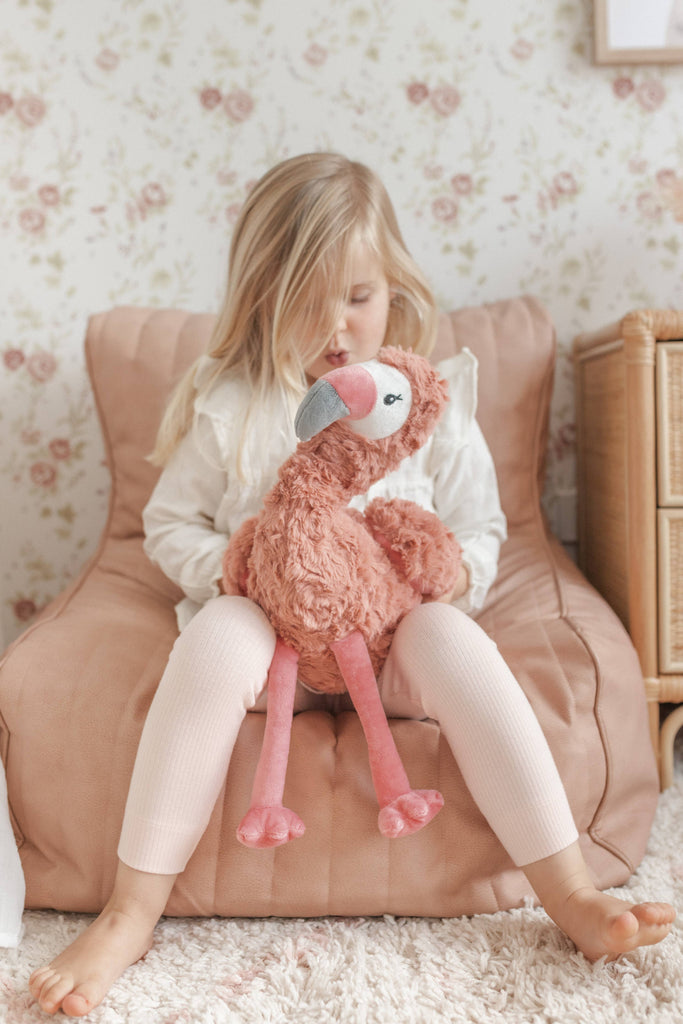 Child sitting on a pink chair holding a plush flamingo toy in a room with floral wallpaper.