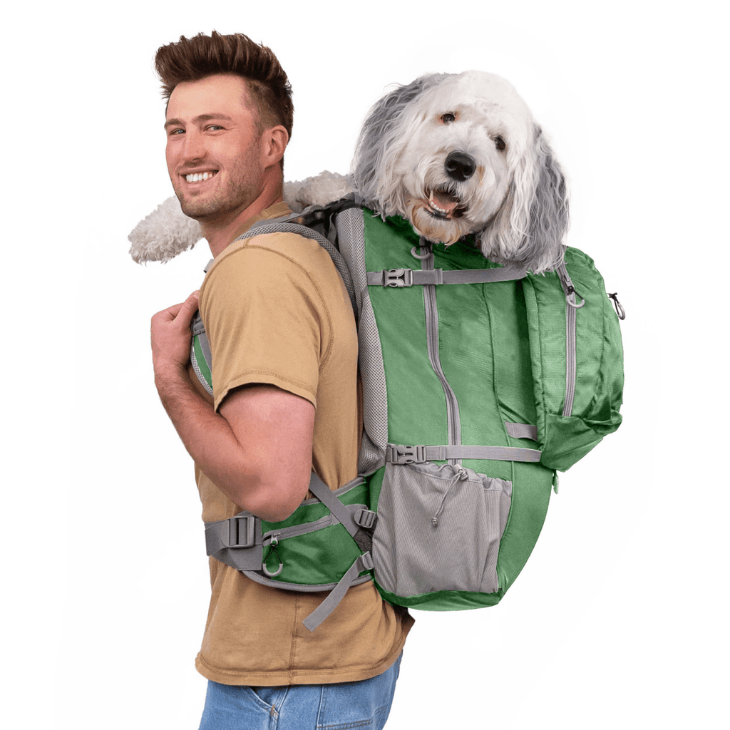 A man wearing a tan t-shirt and blue jeans carries a large green dog backpack with a fluffy white dog inside, photographed against a solid white background.
