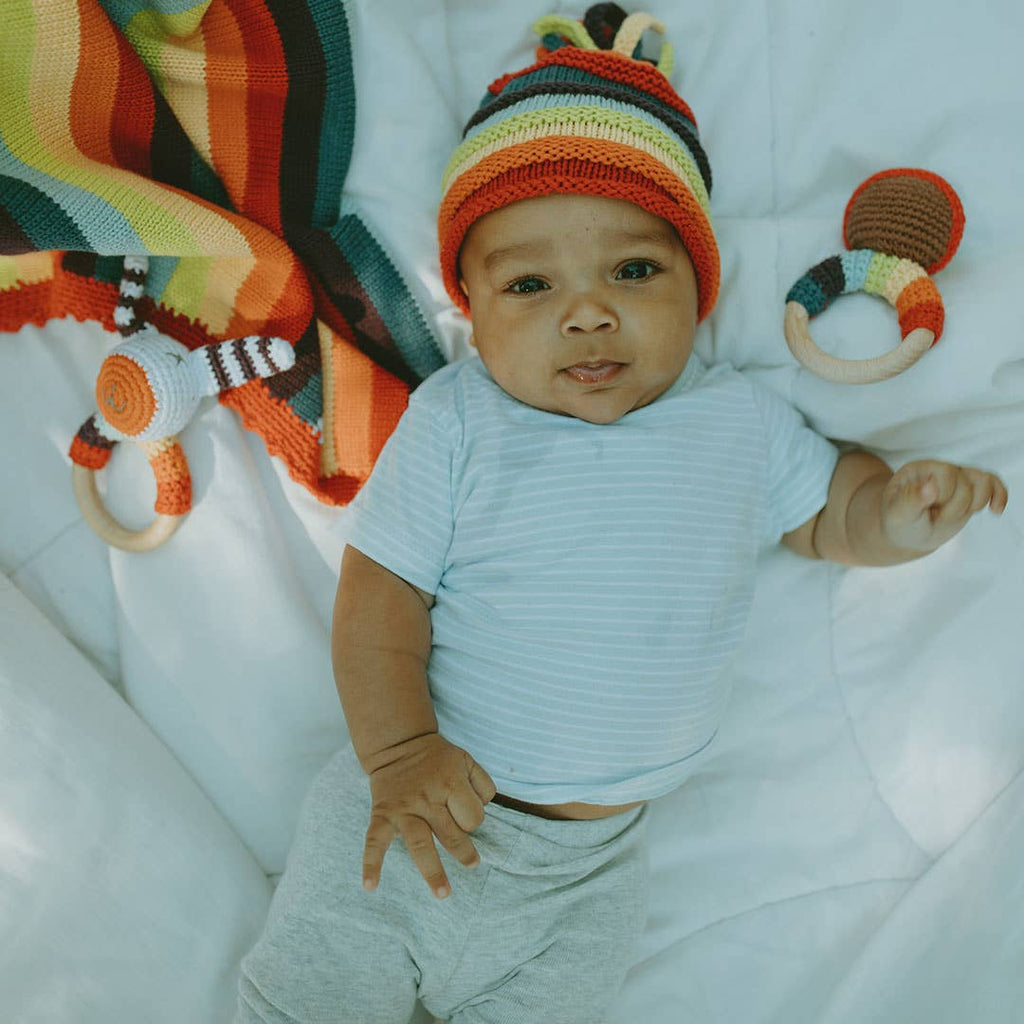 Baby wearing a colorful knit hat with toys around on a white blanket