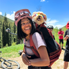 Person with a dog on their back in a forested area with bicycles and people in the background.