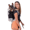 A woman wearing a black and gray dog backpack carrier with a black French Bulldog on her back, set against a solid white background.