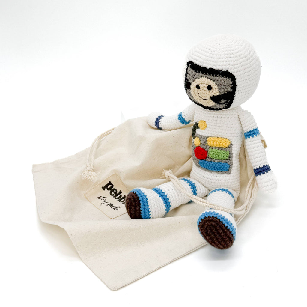 A crocheted astronaut doll in a white spacesuit with blue and brown accents, posed next to a cream-colored canvas drawstring bag with a 'Pebe' brand tag, on a seamless white background.