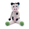 Crocheted cow toy with black spots and green feet on a white background