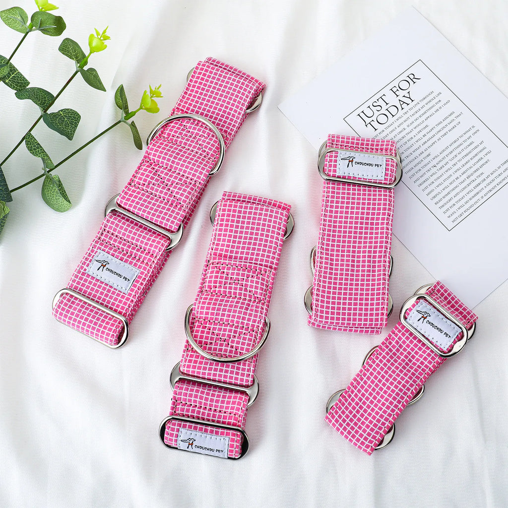 Four pink gingham dog collars with silver metal rings and buckles, arranged on a white fabric background with a decorative green plant and a printed card.
