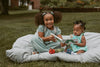 Two young girls sitting on a blanket outdoors, holding toys and smiling.