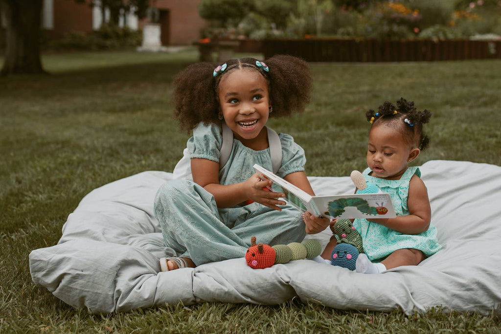 Two young girls sitting on a blanket outdoors, holding toys and smiling.
