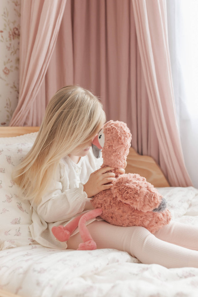 Child holding a pink flamingo plush toy in a bedroom with pink curtains.