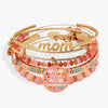 A stack of gold-tone bangles and bracelets with pink and peach beads, floral enamel details, and charms including a large round charm reading 'MOM is just NOW inside out' and a script bangle with 'mom' written across it.
